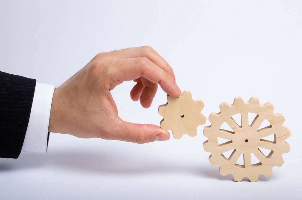 Hand Of Businessman In Suit Holds The Gear To Another Gear Wheel. The Hand Connects Two Round Gears. The Concept Of Business Processes And Ideas, Building A Career And Carrying Ideas. Innovations.