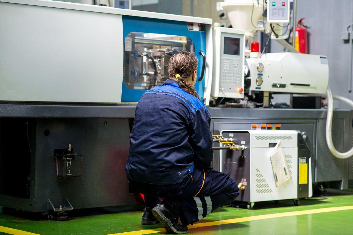 Factory technician performing maintenance and monitoring industrial injection molding equipment inside a manufacturing facility.